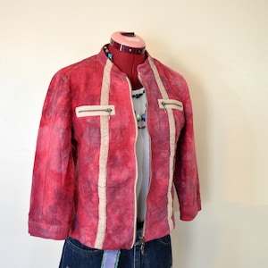 Może przedstawiać: Czerwono-biała kurtka w stylu tie-dye z zamkiem błyskawicznym i białym wykończeniem. Kurtka jest noszona na białym t-shircie i niebieskich dżinsowych spodenkach z fioletowym i zielonym paskiem.