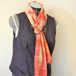 以下が含まれることがあります： オレンジとピンクのタイダイスカーフ、幅20cm、長さ188cmがデニムベストにかけられています。 スカーフはフリンジエッジで、マネキンに展示されています。 テキスト「Scarf #92」が表示されています。