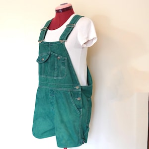 May include: Green denim overall shorts with adjustable straps and multiple pockets. The shorts are displayed on a mannequin with a white t-shirt underneath. The overall shorts have metal hardware and button closures.