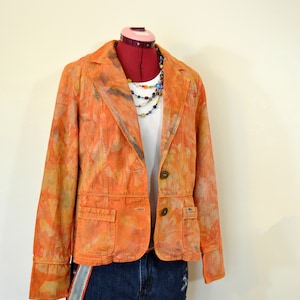 Puede incluir: Una chaqueta naranja con estampado tie-dye, combinada con una camiseta blanca y unos shorts vaqueros. La chaqueta tiene dos bolsillos delanteros y botones marrones. Un collar de cuentas de colores completa el conjunto, expuesto en un maniquí.