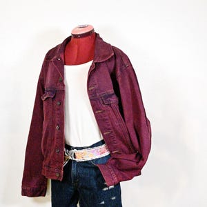 May include: A deep burgundy denim jacket with a collar, button-front closure, and two chest pockets. Underneath is a white ribbed tank top paired with dark blue denim shorts featuring distressed details and a floral patterned fabric belt.