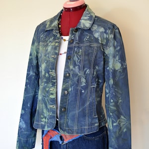 May include: A blue denim jacket with a tie-dye pattern in shades of green and blue. The jacket has a button-up front and long sleeves. It is worn over a white t-shirt and blue denim shorts with a red and blue patterned belt.