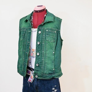 Op de afbeelding: Een groene spijkerweste met zilveren knopen en zakken wordt getoond op een mannequin. De weste wordt gecombineerd met een witte top, een kleurrijke kralenketting, een riem met patroon en een spijkerbroek met distressed details. De outfit is casual en stijlvol.