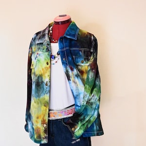 Puede incluir: Una chaqueta vaquera teñida con tintes de azul, verde y amarillo se exhibe en un maniquí. La chaqueta se combina con una camiseta sin mangas blanca, pantalones cortos vaqueros y un cinturón colorido. Un collar de cuentas añade un toque de estilo al conjunto.