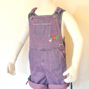Puede incluir: Pantalones cortos de peto vaqueros morados con detalles florales bordados. Los pantalones cortos tienen dobladillos y tirantes ajustables. La prenda se exhibe en un maniquí, sobre una camiseta blanca de manga larga.