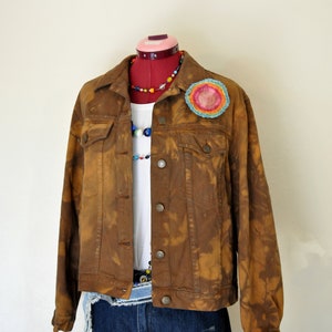 May include: A brown denim jacket with a unique, bleached pattern, worn over a white t-shirt and denim shorts. A colorful, beaded necklace and a decorative, circular patch add pops of color. The outfit is displayed on a mannequin.
