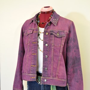 Puede incluir: Una chaqueta vaquera morada con cuello tachonado y una camiseta blanca debajo. La chaqueta tiene un aspecto descolorido y se lleva sobre unos pantalones cortos de mezclilla azules.