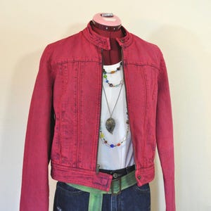 May include: A red denim jacket with a zipper closure, worn over a white t-shirt and blue denim shorts. The outfit includes a green belt and multiple necklaces. The jacket is displayed on a mannequin.