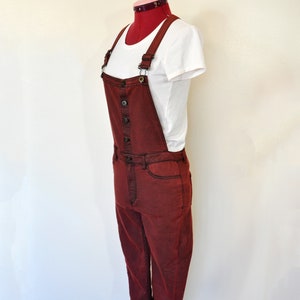 Könnte beinhalten: Eine bordeauxrote Denim-Latzhose mit Knopfverschluss und verstellbaren Trägern. Die Latzhose befindet sich auf einem Mannequin und hat ein weißes T-Shirt darunter.