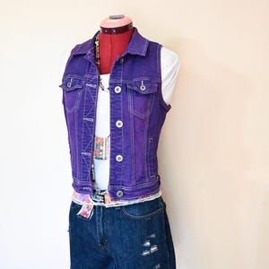 Könnte beinhalten: Eine lila Jeansweste mit weißen Nähten, getragen über einem weißen Oberteil und kombiniert mit Jeansshorts. Das Outfit wird mit einem gemusterten Gürtel und einer Perlenkette ergänzt. Die Shorts haben Distressed-Details.
