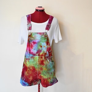 May include: A pair of tie-dye denim overalls with a white t-shirt underneath. The overalls feature a vibrant mix of red, blue, green, and purple hues. The shorts-style overalls have rolled cuffs and adjustable straps.