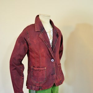 May include: A maroon denim jacket with a single button closure and a pocket on the left side. The jacket is on a mannequin and has a green skirt underneath.