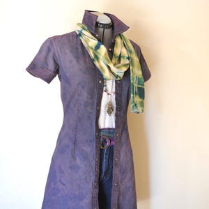 Könnte beinhalten: Ein lila Jeanskleid mit kurzen Ärmeln, Knopfleiste vorne und Kragen. Ein weißes Hemd, eine blaue Jeans und ein gemusterter Schal sind darunter geschichtet. Ein Gürtel und eine Halskette vervollständigen das Outfit, das auf einer Schaufensterpuppe präsentiert wird.