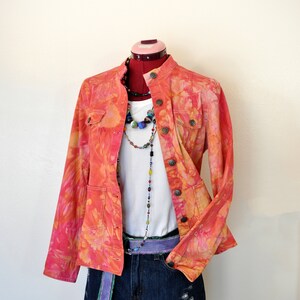 May include: A tie-dye denim jacket with a white t-shirt and blue denim shorts. The jacket has a mandarin collar and a button closure. The shorts have a distressed look.