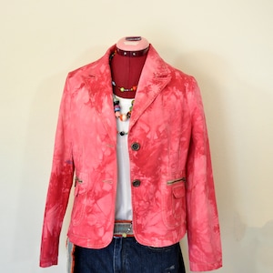 May include: A pink tie-dye blazer with two front pockets and a two-button closure. The blazer is paired with a white top, denim shorts, and a colorful beaded necklace. The outfit is displayed on a red mannequin.