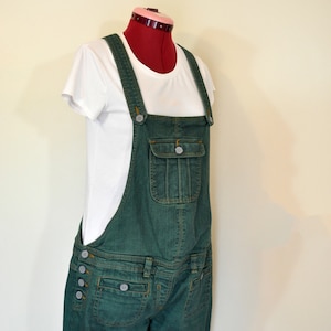 May include: Green denim dungarees with adjustable straps and button fastenings. The dungarees have a bib pocket, side pockets, and back pockets. A white t-shirt is worn underneath.