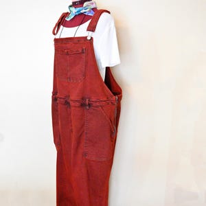 Könnte beinhalten: Rote Jeans-Latzhose mit mehreren Taschen und verstellbaren Trägern, präsentiert auf einer Schaufensterpuppe. Darunter wird ein weißes T-Shirt getragen. Ein farbenfrohes Halstuch ist um den Hals der Schaufensterpuppe gebunden. Die Latzhose hat eine lockere Passform und ein verwaschenes Aussehen.