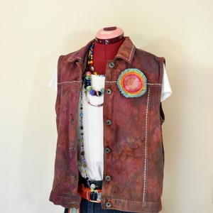 May include: A burgundy vest with white stitching, a colorful circular patch, and a white t-shirt. The vest is paired with denim shorts and a beaded necklace. A red belt with a striped accent is also visible.