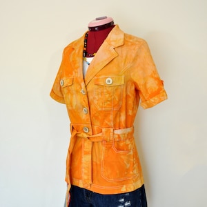 Puede incluir: Chaqueta naranja teñida con tinte anudado con mangas cortas, cinturón y botones blancos grandes. La chaqueta tiene dos bolsillos en el pecho y se combina con pantalones cortos vaqueros azul oscuro. Se ve un collar de cuentas de colores.