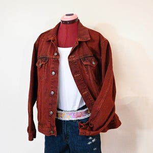 May include: A brick-red denim jacket with silver buttons, worn over a white ribbed tank top and distressed denim shorts. A colorful patterned belt adds a pop of color to the outfit. The ensemble is displayed on a mannequin.
