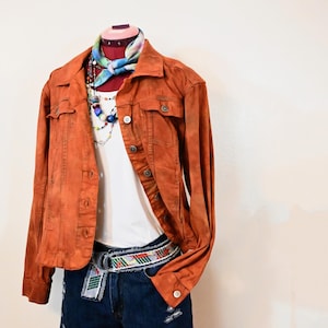 Puede incluir: Una chaqueta vaquera de color óxido con un top blanco sin mangas, un collar de cuentas de colores y un pañuelo de bandana teñido anudado. La chaqueta se lleva sobre unos pantalones cortos de mezclilla azul oscuro con un cinturón tejido de colores.