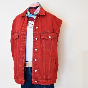 Puede incluir: Un chaleco vaquero rojo con botones blancos, usado sobre una camiseta blanca y pantalones cortos vaqueros. El conjunto se complementa con una bufanda colorida, un collar de cuentas y un cinturón estampado. El chaleco tiene dos bolsillos delanteros.