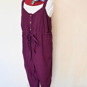 Könnte beinhalten: Ein tiefvioletter Jumpsuit mit Knopfleiste vorne und verstellbarem Taillenband. Der Jumpsuit hat breite Träger und eine lockere Passform. Darunter wird ein weißes Oberteil getragen. Ein farbenfrohes Halstuch ist um den Hals der Schaufensterpuppe drapiert.