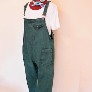 Op de afbeelding: Groene denim overall met grote voorzakken en verstelbare bandjes. Er wordt een wit t-shirt onder gedragen en een kleurrijke sjaal is om de nek van de mannequin gedrapeerd. De overall heeft een relaxte pasvorm en een klassiek ontwerp.