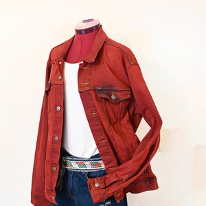 May include: A red denim jacket with a white t-shirt underneath and blue denim shorts with a colorful woven belt.