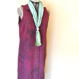 May include: A sleeveless, button-down dress in a deep burgundy colour, featuring two front pockets. A light teal scarf is knotted around the neck of a mannequin. The dress has a faded, textured appearance.