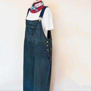 Op de afbeelding: Een lange denim overall jurk met een voorzak en goudkleurige knopen. De jurk wordt gecombineerd met een wit t-shirt en een kleurrijke sjaal. De overall jurk wordt gepresenteerd op een mannequin.