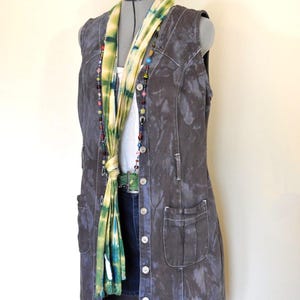 May include: A long, sleeveless denim vest in a dark grey wash, with button fastenings and front pockets. A tie-dye scarf in shades of green and yellow is draped over the vest, along with a beaded necklace. The vest is displayed on a mannequin.