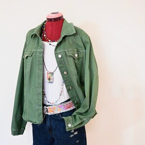May include: An olive green denim jacket worn over a white ribbed tank top and dark blue distressed denim shorts. The outfit includes a multi-colored beaded necklace with a rectangular pendant and a wide belt featuring a floral pattern.