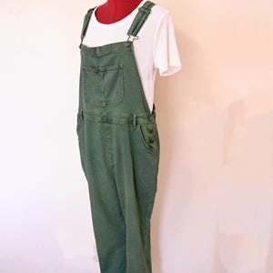 Può includere: Salopette in denim verde con spalline regolabili e più tasche. Sotto è indossata una maglietta bianca a maniche corte. La salopette è esposta su un manichino con collo rosso.