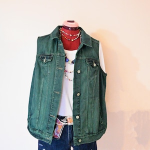 Op de afbeelding: Een groene spijkerweste over een witte tanktop, gecombineerd met een spijkerbroek en een riem met patroon. Een rode mannequinhoofd draagt ​​gelaagde kettingen en een choker. De outfit wordt gepresenteerd op een rode standaard.