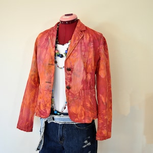 May include: A vibrant orange and red tie-dye blazer with three brown buttons, paired with a white top and denim shorts. A colorful beaded necklace and a denim belt complete the outfit, displayed on a red dress form.