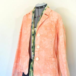 Könnte beinhalten: Ein pfirsichfarbener Blazer mit Batikmuster, drei weißen Knöpfen und zwei Vordertaschen. Ein grüner und blauer Schal und eine Halskette sind sichtbar. Der Blazer wird auf einer Schaufensterpuppe präsentiert.