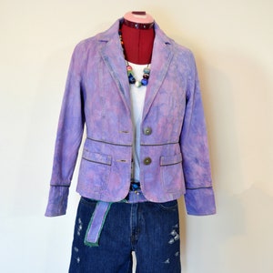 May include: A lilac blazer with a subtle tie-dye effect, paired with a white top and denim shorts. The outfit includes a colorful beaded necklace and a denim belt with a green accent. The blazer has two front pockets and dark brown trim.
