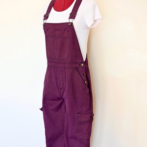 May include: A pair of deep purple overalls with adjustable straps and multiple pockets. The overalls are displayed on a mannequin wearing a white t-shirt. The garment has a classic, casual style, suitable for everyday wear.