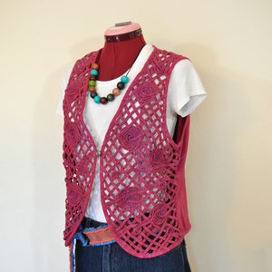 May include: A pink crocheted vest with floral details, worn over a white t-shirt and blue denim shorts. The vest has a criss-cross pattern and is accented with a multi-colored beaded necklace.
