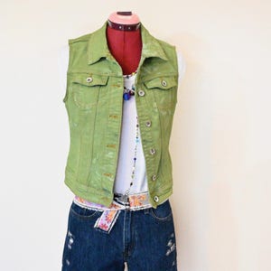 May include: A green denim vest with button closures is displayed on a mannequin. The vest is paired with a white top, distressed denim shorts, and a floral belt. A colorful beaded necklace adds a pop of color to the outfit.