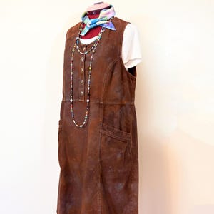Puede incluir: Un vestido de cuero marrón sin mangas con cierre de botones en la parte delantera y dos bolsillos. El vestido está en un maniquí y tiene una bufanda colorida y un collar de cuentas.