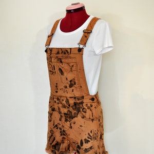 May include: Brown denim overall shorts with a black tie-dye pattern. The shorts have a frayed hem and are paired with a white t-shirt. The overall straps are adjustable and feature black metal hardware.