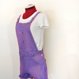 Puede incluir: Un conjunto de peto corto de mezclilla morada exhibido en un maniquí. El pantalón corto tiene un dobladillo deshilachado y se combina con una camiseta blanca. El peto presenta botones y herrajes de color cobre.