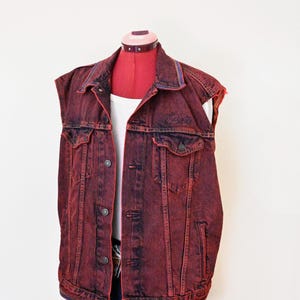May include: A sleeveless denim vest in a deep red acid-wash finish featuring a classic collar, a front button closure, two chest pockets with buttoned flaps, and two side pockets. The vest is displayed on a mannequin over a white tank top.