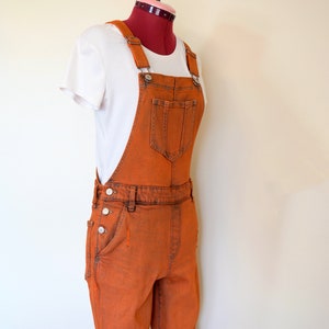 Puede incluir: Mono vaquero naranja con tirantes ajustables, un bolsillo en el peto y cierres laterales con botones. El mono se combina con una camiseta blanca. El conjunto se exhibe en un maniquí.