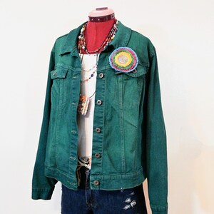 Op de afbeelding: Een groene spijkerjas met een kleurrijke gehaakte bloemspeld, gelaagd over een witte top en een spijkerbroek. De mannequin is versierd met kralenkettingen. De shorts hebben distressed details.