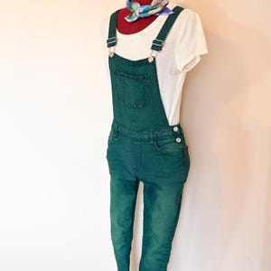 Può includere: Una salopette di jeans verde con una maglietta bianca e una sciarpa colorata a motivi. La salopette ha bretelle regolabili e una tasca anteriore.