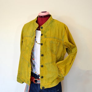 Op de afbeelding: Een geel denim jack met een mandarijnkraag en knoopsluiting. Het jack heeft twee voorzakken en een wit shirt eronder. Het jack wordt gedragen over een blauwe denim short met een kleurrijke riem.