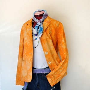May include: An orange blazer with a tie-dye pattern, paired with a white top, denim shorts, and a purple belt. A colorful scarf and beaded necklace add accents to the outfit. The outfit is displayed on a mannequin.